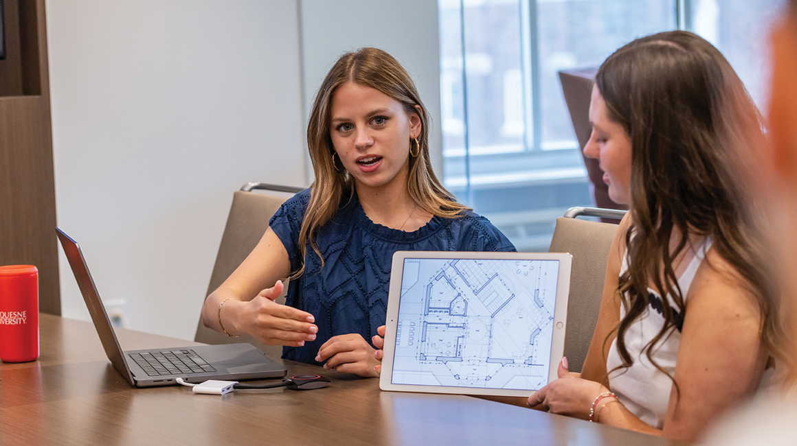 Students sit at a table and discuss renderings on a tablet.