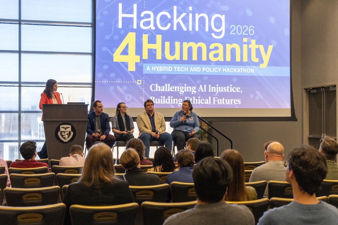 1 moderator stands at lectern and 4 panelists sit onstage for an expert discussion of AI injustice