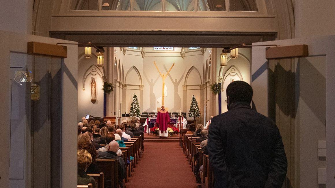 A man stands at in the narthex of the Chapel, looking in as the Mass is celebrated and a priest delivers the homily to the assembly.